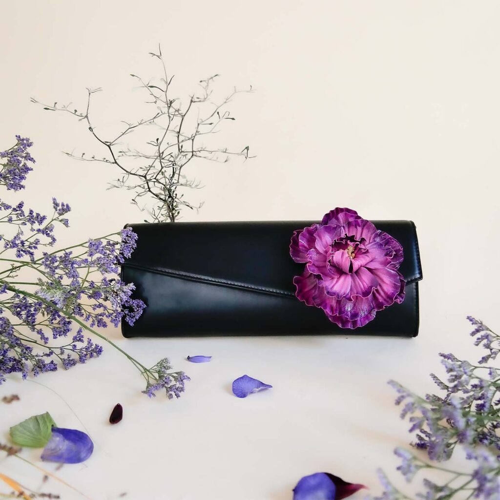 Clutch aus schwarzem Leder mit abnehmbarer lila Pfingstrose aus Leder, vor hellem Hintergrund mit Blüttenblättern und Zweigen