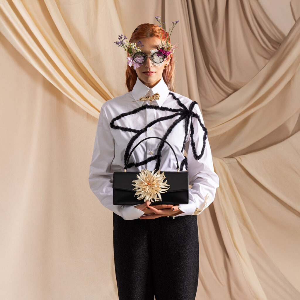 Klassische Handtasche aus schwarzem Leder mit wechselbarer Blume aus Leder, gehalten von Model in weißer Bluse