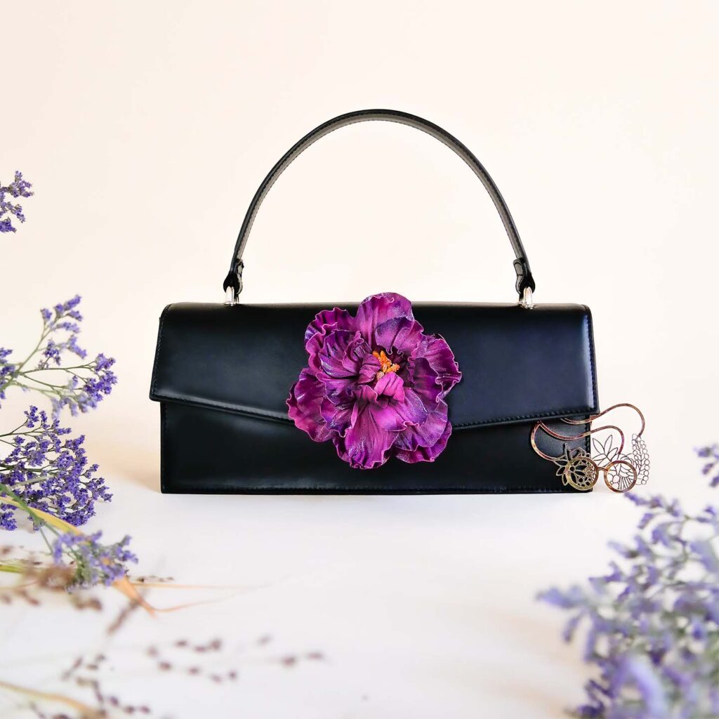 Wandelbare Handtasche aus schwarzem Leder mit abnehmbarer Lederblume in lila, vor weißem Hintergrund mit Zweigen