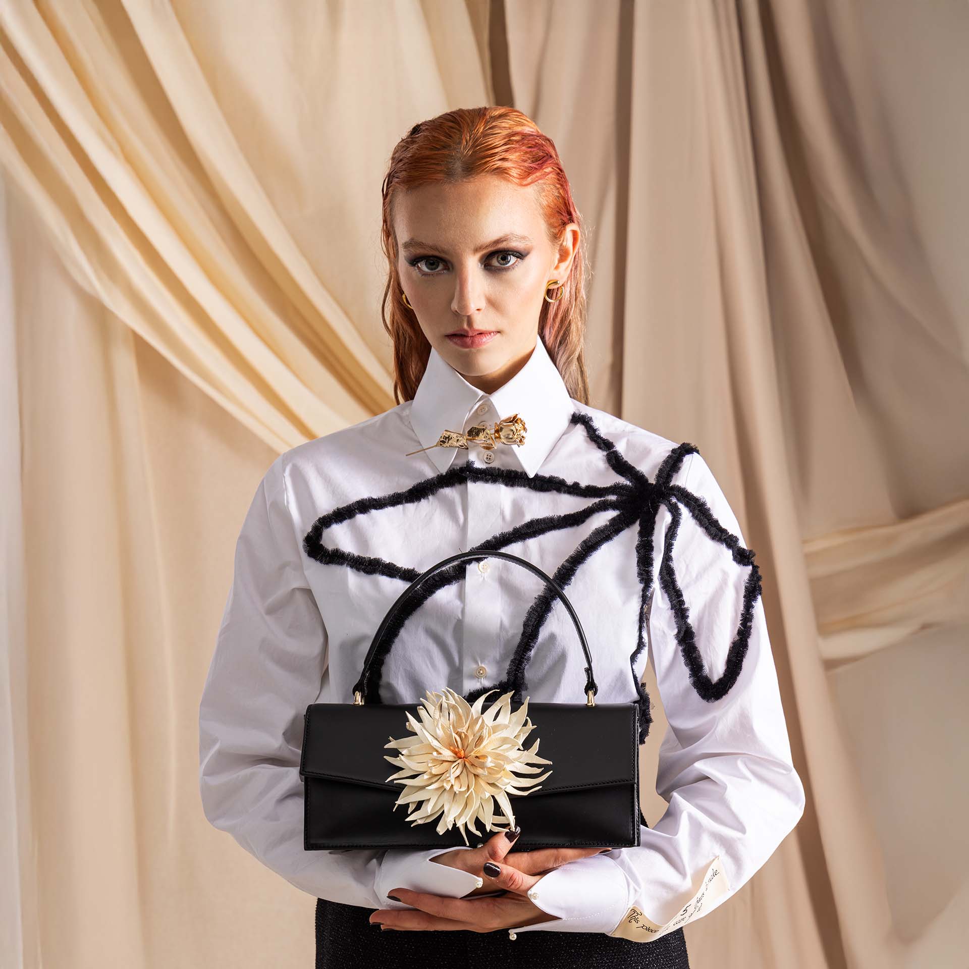 Handtasche klassischer Form aus schwarzem Leder in mit weißer, abnehmbarer Dahlienblüte, getragen von junger Frau mit weißer Bluse