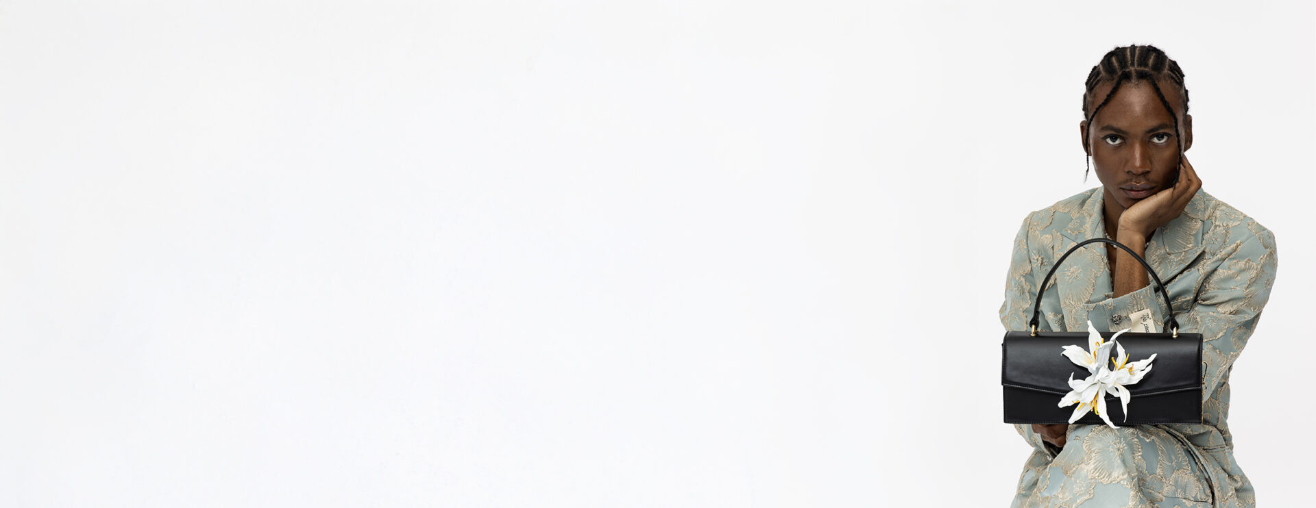 Premium Lederhandtasche Modell "Gaia" mit abnehmbarer weißer Lederblume "Lily", gehalten von sitzendem Model in Anzug mit Blumenmuster