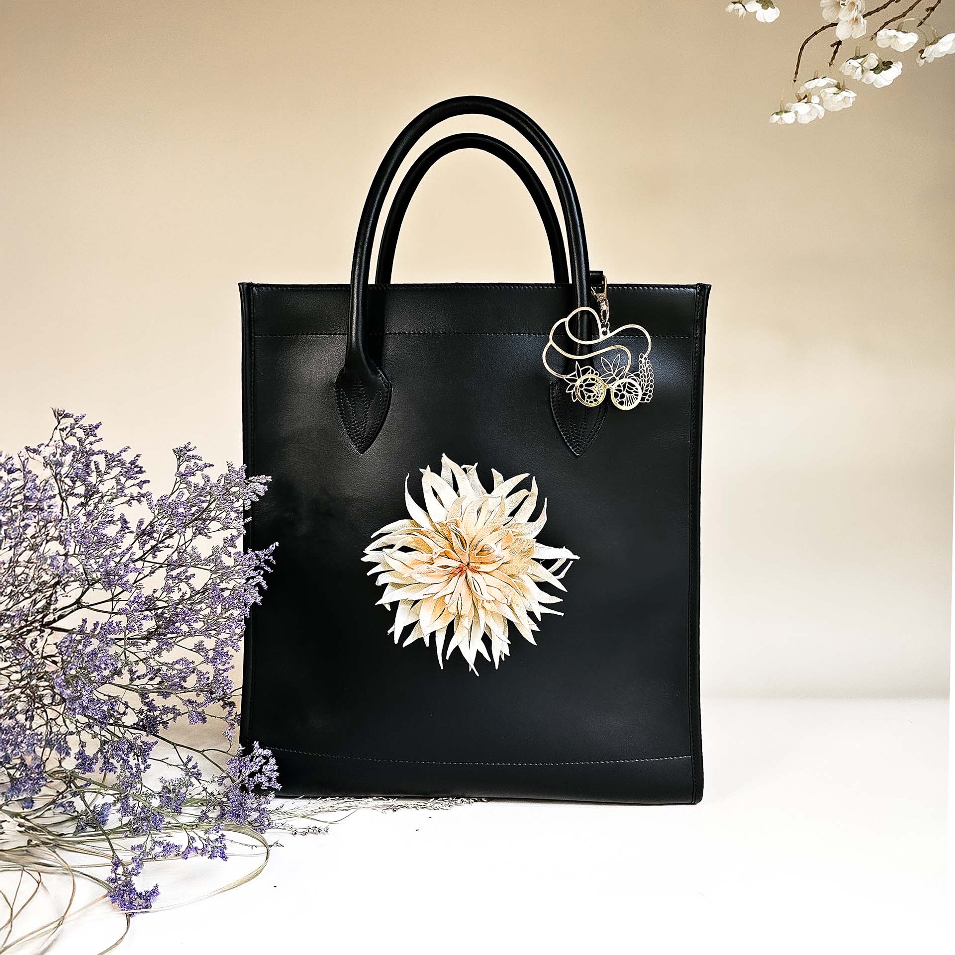Tote-Bag aus schwarzem Leder mit abnehmbarer weißer Dahlie und goldenem Metallelement in hellem Hintergrund mit dekorativen Zweigen